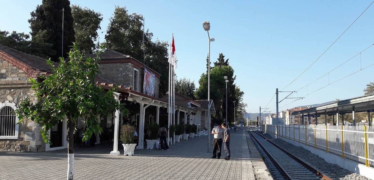 La estación de tren de Selçuk: el edificio antiguo de piedra con tejados de teja y porche cubierto, una bandera turca, el andén y la vía electrificada que se aleja hacia la derecha
