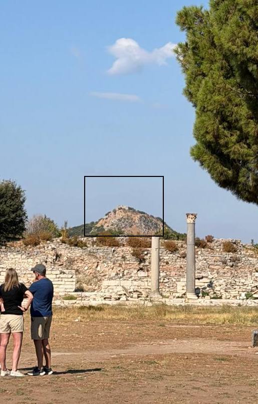 Una colina rocosa al oeste de las ruinas de Éfeso con una pequeña torre de piedra en la cima, marcada con un recuadro negro, vista tras un muro en ruinas y dos columnas, con una pareja de visitantes de espaldas